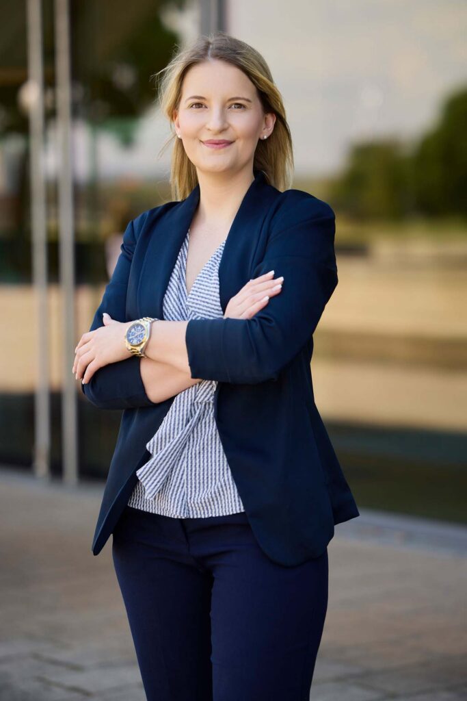 Eine Frau mit blondem Haar steht im Freien und lächelt selbstbewusst mit verschränkten Armen - bereit, Baufinanzierung Beratung anzubieten. Sie trägt einen marineblauen Blazer, eine gestreifte Bluse und eine Uhr. Im Hintergrund sind ein modernes Gebäude und Bäume unscharf zu erkennen.