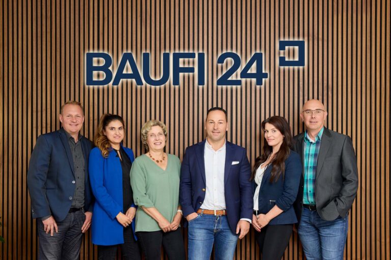 Vor einer Holzlattenwand mit dem BAUFI24-Logo stehen sechs professionell gekleidete Personen, die für eine kompetente Baufinanzierungsberatung stehen und freundlich lächelnd in die Kamera posieren.