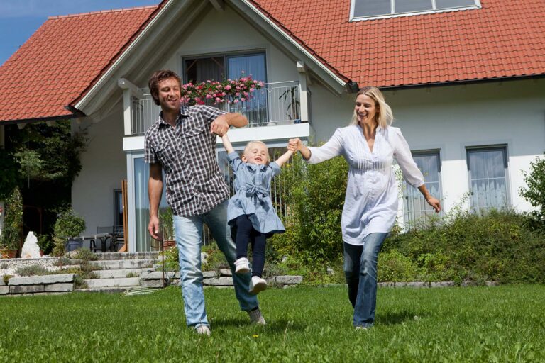 Ein lächelnder Mann und eine lächelnde Frau schwingen ein junges Mädchen auf ihren Armen, während sie auf dem grünen Rasen vor einem modernen Haus spazieren gehen. Sie genießen die Zeit mit ihrer Familie, nachdem sie sich mit Hilfe von baufinanzierung beratung ihr Traumhaus gesichert haben.