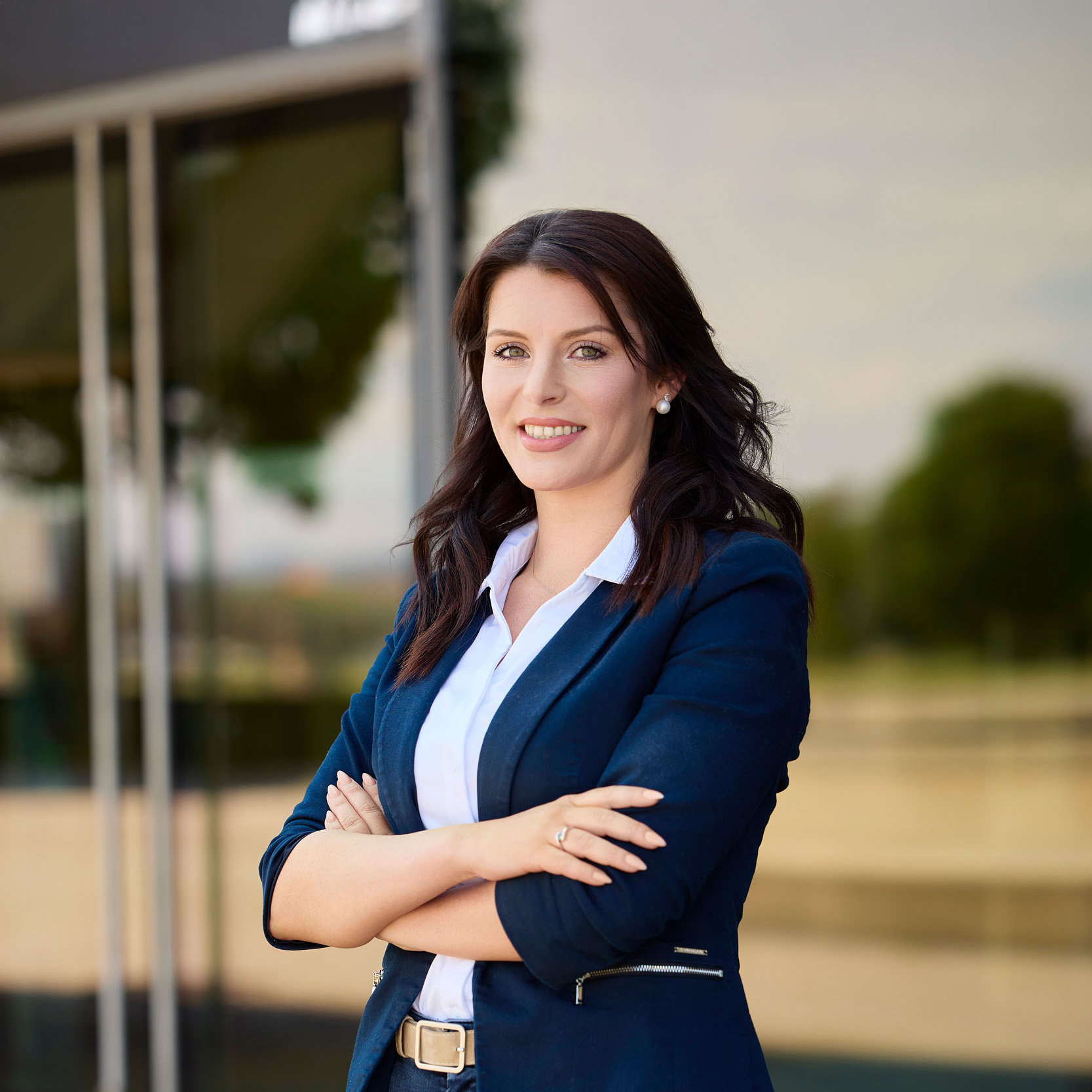 Eine Frau mit langen dunklen Haaren, die einen blauen Blazer über einem weißen Hemd trägt, steht draußen mit verschränkten Armen und lächelt selbstbewusst in die Kamera - eine Expertin, die bereit ist, Baufinanzierungsberatung in Dresden oder Leipzig anzubieten. Glasfenster und eine verschwommene Außenlandschaft bilden den Hintergrund.