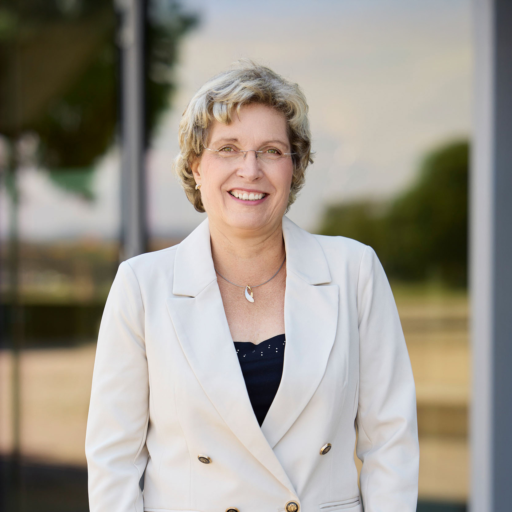 Eine Frau mit kurzen blonden Haaren und Brille lächelt, während sie im Freien steht, vielleicht nach einer baufinanzierung beratung. Sie trägt einen weißen Blazer über einem dunklen Oberteil, mit verschwommenen Bäumen und Gebäuden im Hintergrund.