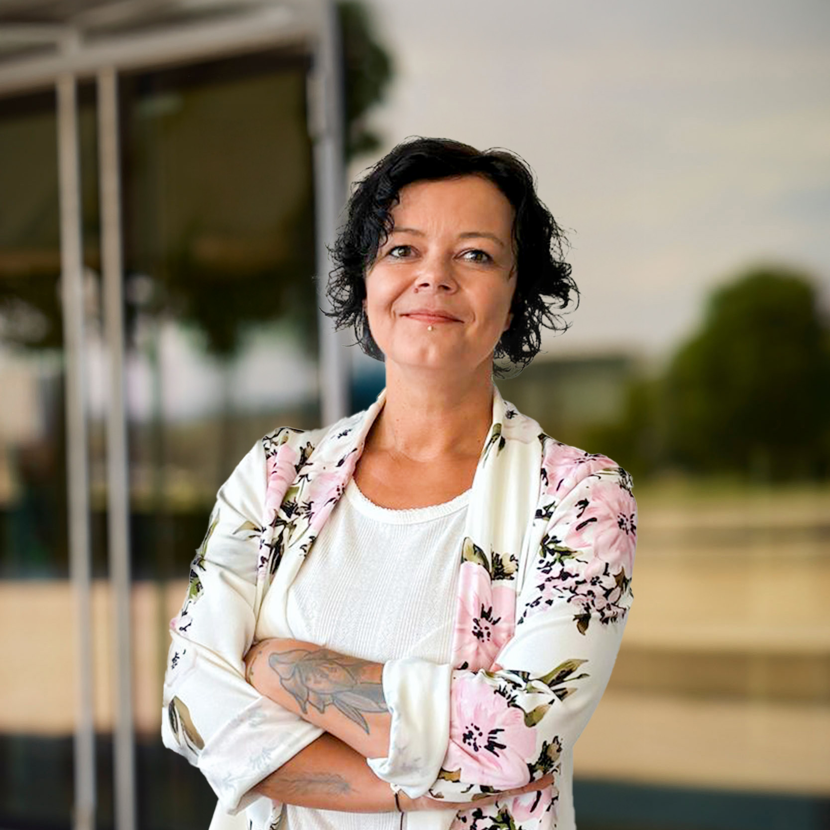 Eine Frau mit kurzen lockigen dunklen Haaren, die ein weißes Hemd und einen geblümten Blazer trägt, steht mit verschränkten Armen und lächelt selbstbewusst - perfekt, um Baufinanzierung Beratung in einer modernen Büroumgebung mit Glas und Grün im Hintergrund zu repräsentieren.