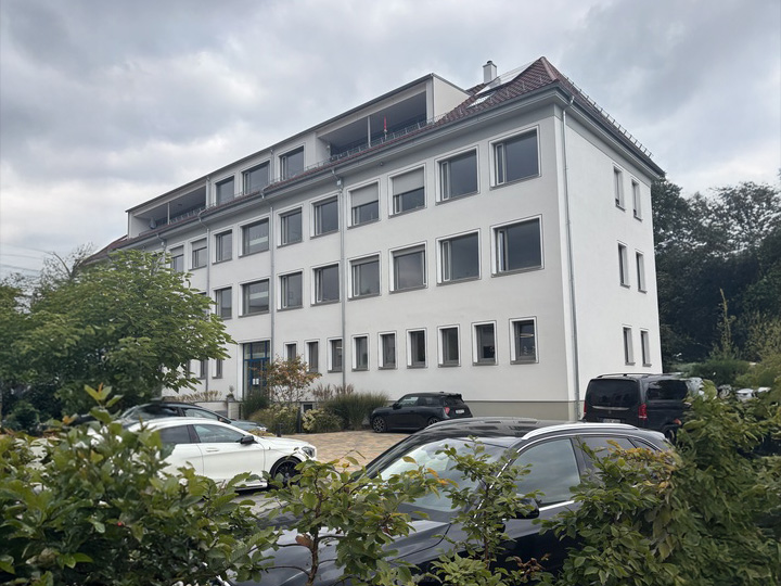 Ein weißes, dreistöckiges Gebäude mit Ziegeldach und vielen Fenstern ist umgeben von Grünflächen und parkenden Autos - ein passender Ort für die baufinanzierung beratung in Leipzig. Der Himmel ist bedeckt.