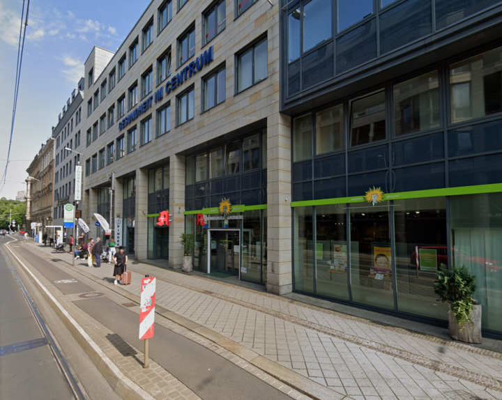 Ein modernes, mehrstöckiges Gebäude mit Glas- und Steinfassade, in dem eine Apotheke und andere Geschäfte untergebracht sind. Menschen warten in der Nähe der Straßenbahnlinie - perfekt gelegen für diejenigen, die eine baufinanzierung Leipzig oder eine kompetente baufinanzierung beratung suchen.