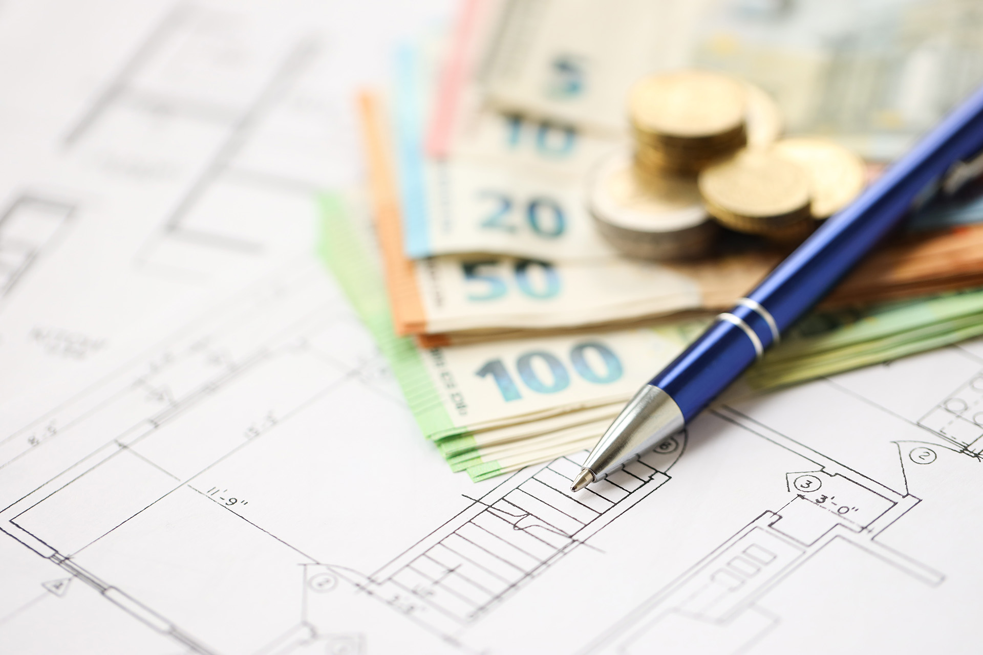 Eine Nahaufnahme von architektonischen Entwürfen mit einem Stift, Euro-Banknoten und Münzstapeln, die die finanzielle Planung oder Budgetierung eines Bauprojekts symbolisieren - ideal für diejenigen, die Baufinanzierung Beratung in Leipzig oder Dresden suchen.