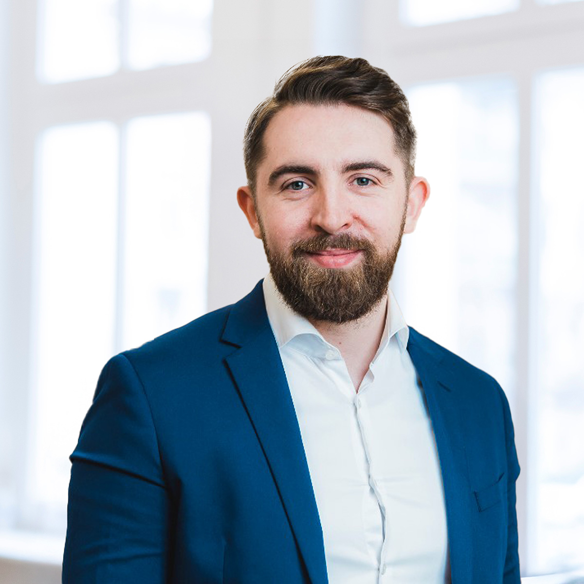 Ein Mann mit kurzen braunen Haaren und Bart, der ein weißes Hemd und einen blauen Blazer trägt, steht lächelnd vor den hellen, verschwommenen Fenstern.