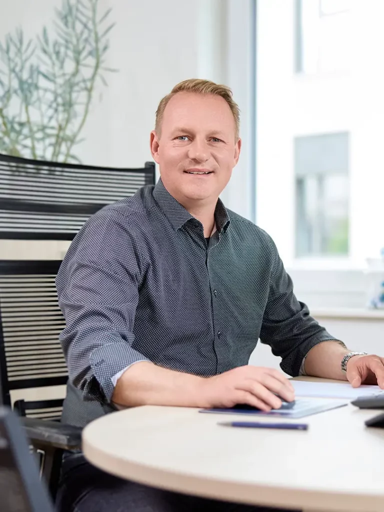 Ein Mann mit kurzem blondem Haar sitzt an einem Schreibtisch und lächelt in die Kamera. Er trägt ein dunkel gemustertes Hemd und hat eine Hand auf einem Tablet, die andere ruht auf dem Tisch, ein Stift liegt daneben. Im Hintergrund sind ein Fenster und eine Pflanze zu sehen.
